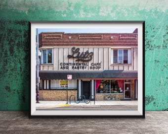 May include: Framed print of Lutz Continental Cafe and Pastry Shop. The building has a brick facade, a black awning, and a sign that reads "Lutz Continental Cafe and Pastry Shop". The shop has large windows displaying pastries and cakes.