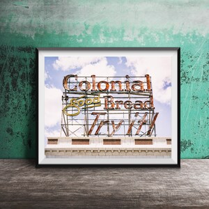 Può includere: Stampa incorniciata di un'insegna vintage Colonial Good Bread contro un cielo blu con nuvole. L'insegna presenta le parole "Colonial Good Bread" e "Try It" in un font retrò, con un aspetto usurato e arrugginito.