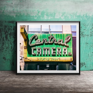 Könnte beinhalten: Ein vintage grünes und gelbes Neon-Schild mit dem Text "Central Camera. Since 1899." Das Schild ist an einem Gebäude mit einer Backsteinfassade angebracht.