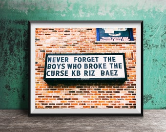 May include: A black and white sign with the text "NEVER FORGET THE BOYS WHO BROKE THE CURSE KB RIZ BAEZ" on a brick wall. The sign is mounted on a white way.