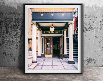 May include: A framed photographic print of the Pizzeria Due entrance, featuring a dark facade with gold lettering, white fluted columns with gold capitals, a hanging lantern, and a wooden door, all set against a light-colored tiled entryway.