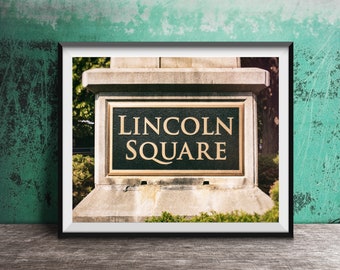 May include: A framed wall art print featuring a photograph of a stone monument base with a dark green rectangular plaque. The plaque has a gold border and the text LINCOLN SQUARE in gold capital letters. The monument is set against green trees.