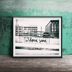 May include: Black and white photograph of a city street with the words "I love you" spray painted on a concrete wall.