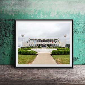 Może przedstawiać: Oprawiona fotografia budynku muzeum Indianapolis Motor Speedway, z beżową fasadą z napisem Indianapolis Motor Speedway Racing Capital of the World, centralnym wejściem, zielenią oraz betonową ścieżką prowadzącą do budynku.