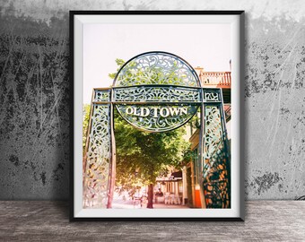 May include: A framed wall art print featuring a decorative dark metal archway with intricate scrollwork patterns. The center of the arch displays the text OLD TOWN in bold white lettering, positioned above a sunlit street scene with green trees.