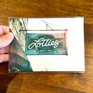 Pode incluir: Uma placa de madeira desgastada com o texto "Lotties Since 1934" em letras douradas. A placa est&aacute; pendurada em uma corrente e tem um esquema de pintura vermelho, branco e azul.