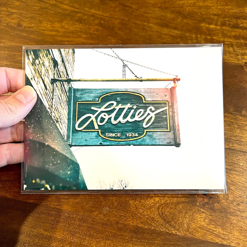Pode incluir: Uma placa de madeira desgastada com o texto "Lotties Since 1934" em letras douradas. A placa est&aacute; pendurada em uma corrente e tem um esquema de pintura vermelho, branco e azul.