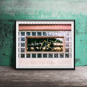 May include: Framed photograph of a building facade with a window featuring a neon sign of a dog. The window is surrounded by glass blocks and a wooden frame. The background is a textured teal wall.