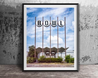 May include: A framed print of a vintage bowling alley sign. The sign spells out "BOWL" in large, white letters on circular black backgrounds. The image is set against a blue sky with clouds, and the building's architecture is visible below.