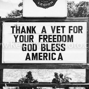 May include: A monochrome sign with the message "THANK A VET FOR YOUR FREEDOM GOD BLESS AMERICA A". The sign is mounted on a wooden frame, with a Legion emblem at the top. The background includes trees and a road.