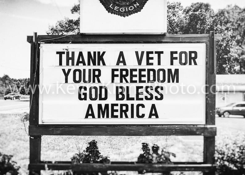 May include: A monochrome sign with the message "THANK A VET FOR YOUR FREEDOM GOD BLESS AMERICA A". The sign is mounted on a wooden frame, with a Legion emblem at the top. The background includes trees and a road.