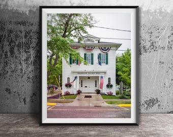 May include: Framed print of Saugatuck Village Hall, a white building with green shutters and American flags. The building has a symmetrical design with a central entrance and a sign that reads "Saugatuck Village Hall". The print is displayed on a wooden surface.