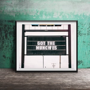 以下が含まれることがあります： 「GOT THE MUNCHES」と白い大文字で書かれた看板が写った額入りの写真。看板は黒色で、文字用のスロットのグリッドがあります。背景は風化した緑色の壁と木製の表面です。