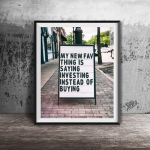 May include: A black and white sign with the text "My new fav thing is saying investing instead of buying" on a brick sidewalk in front of a building.