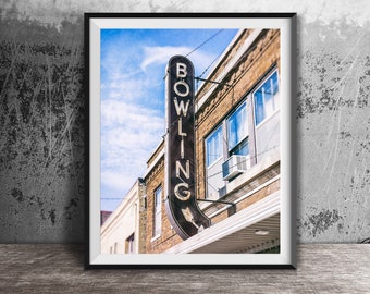 May include: Framed photograph of a vintage bowling alley sign. The sign, with the word "BOWLING" in vertical, illuminated letters, hangs against a brick building. The sky is blue with scattered clouds. The frame is black, and the image is set on a wooden surface.