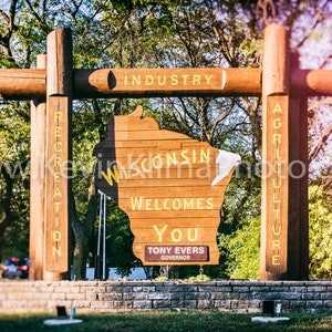 May include: A wooden sign shaped like the state of Wisconsin welcomes visitors with the text "Wisconsin Welcomes You" and the name of the governor, Tony Evers. The sign is surrounded by trees and a stone wall.