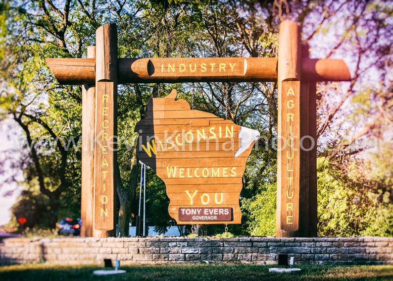 May include: A wooden sign shaped like the state of Wisconsin welcomes visitors with the text "Wisconsin Welcomes You" and the name of the governor, Tony Evers. The sign is surrounded by trees and a stone wall.