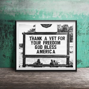 May include: Monochrome framed sign displaying the message "THANK A VET FOR YOUR FREEDOM GOD BLESS AMERICA". The sign is set against a blurred outdoor backdrop, with the word "LEGION" visible at the top. The image is framed in black.