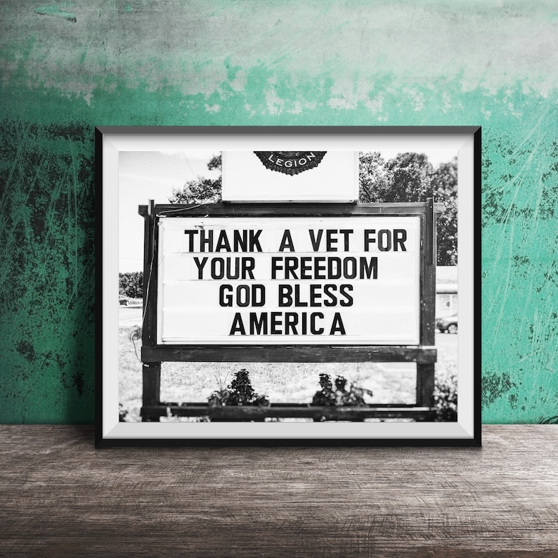 May include: Monochrome framed sign displaying the message "THANK A VET FOR YOUR FREEDOM GOD BLESS AMERICA". The sign is set against a blurred outdoor backdrop, with the word "LEGION" visible at the top. The image is framed in black.