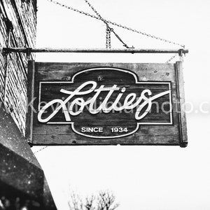 Pode incluir: Fotografia a preto e branco de uma placa de madeira pendurada num edif&iacute;cio. A placa diz "Lotties Since 1934".