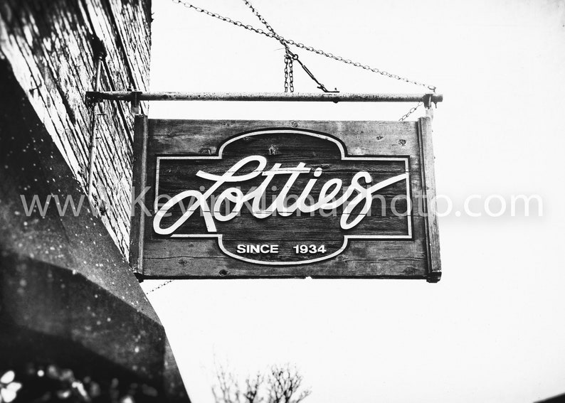 Pode incluir: Fotografia a preto e branco de uma placa de madeira pendurada num edif&iacute;cio. A placa diz "Lotties Since 1934".