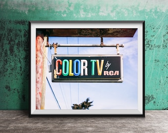 May include: A rectangular outdoor sign with a black background featuring the multi-colored text COLOR TV by RCA in a retro font. The sign is mounted on a metal pole against a blue sky, showing signs of weathering and age on the frame and support.