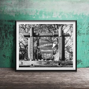 May include: Black and white photo of a wooden sign welcoming visitors to Wisconsin. The sign features the state's outline and the words "Wisconsin Welcomes You" with the name of the governor, Tony Evers, below. The sign is flanked by two wooden posts with the words "Recreation", "Industry", and "Agriculture" written on them.