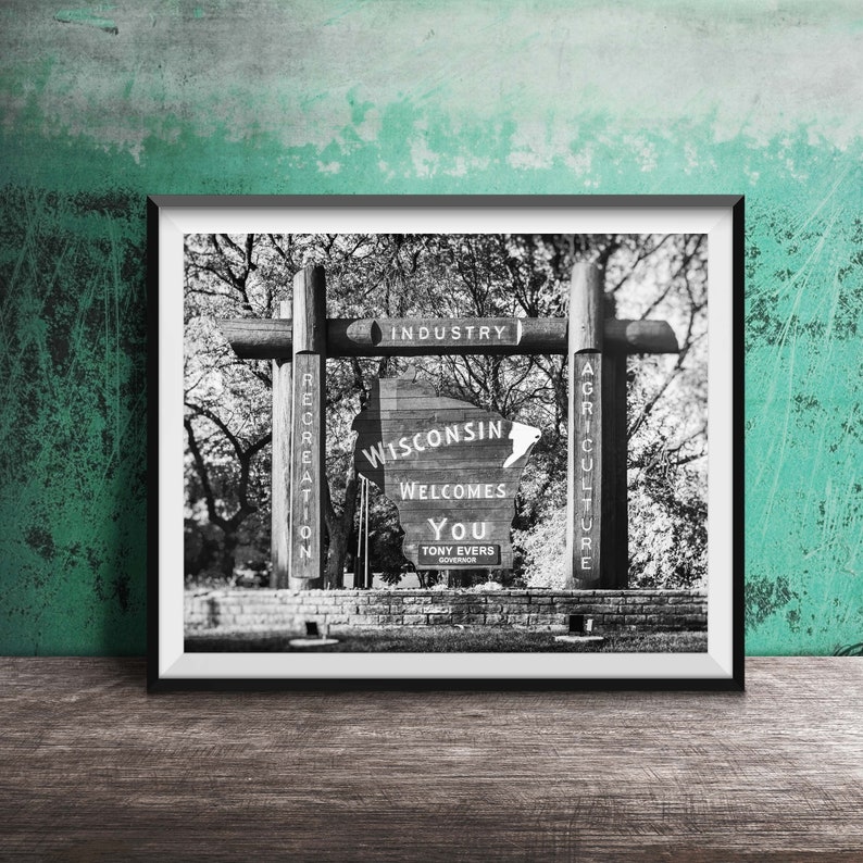 May include: Black and white photo of a wooden sign welcoming visitors to Wisconsin. The sign features the state's outline and the words "Wisconsin Welcomes You" with the name of the governor, Tony Evers, below. The sign is flanked by two wooden posts with the words "Recreation", "Industry", and "Agriculture" written on them.