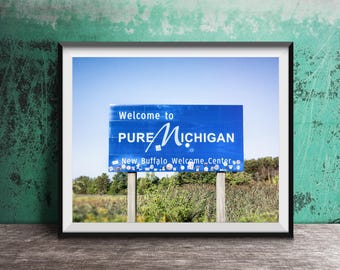 May include: Framed print of a blue sign that reads "Welcome to Pure Michigan" with "New Buffalo Welcome Center" below. The sign is set against a blue sky and a backdrop of green foliage.