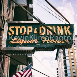 Op de afbeelding: Een vintage neon-bord met de woorden "STOP & DRINK Liquor House" hangend aan een ketting boven een bakstenen gebouw. Het bord is verlicht met gele neonlichten en heeft een groene achtergrond.