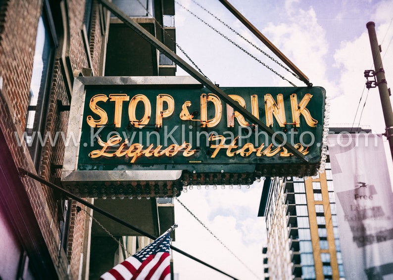 Op de afbeelding: Een vintage neon-bord met de woorden "STOP & DRINK Liquor House" hangend aan een ketting boven een bakstenen gebouw. Het bord is verlicht met gele neonlichten en heeft een groene achtergrond.