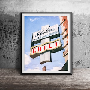 Może przedstawiać: Oprawiony wydruk przedstawiający szyld w stylu vintage dla Skyline Chili. Szyld ma białe tło z napisem Skyline niebieską czcionką nad grafiką panoramy miasta oraz napisem CHILI czerwonymi literami na dolnej sekcji z widocznymi żarówkami.