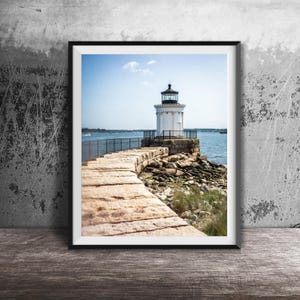 May include: A framed photograph of a white lighthouse with a black lantern room, situated at the end of a stone breakwater. The structure is surrounded by blue water under a clear sky, with rocky terrain visible along the base of the lighthouse.
