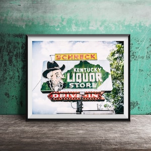 Puede incluir: Un letrero de neón vintage para una tienda de licores de Kentucky con las palabras "Schreck Kentucky Liquor Store" y "Drive In" en letras rojas y blancas. El letrero presenta a un hombre de dibujos animados con un sombrero de vaquero y una chaqueta verde.