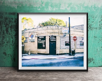 May include: A framed photograph of a corner building labeled Carol's Pub, featuring brick walls, a dark entrance door, and signage for Old Style beer, pool, darts, and sandwiches. A stop sign and a two-way traffic sign are visible on the street corner.