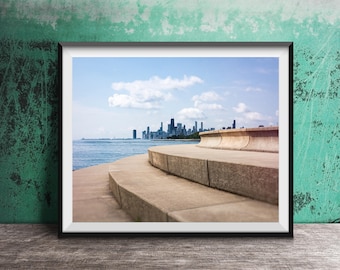 May include: A framed photograph featuring a perspective view of curved concrete steps leading toward a body of water. In the distance, a city skyline with various skyscrapers is visible under a bright blue sky with scattered white clouds.