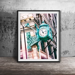 Op de afbeelding: Een vintage-stijl klok met Romeinse cijfers is gemonteerd op een gevel van een gebouw. De klok is ingelijst in een zwart frame en omgeven door sierlijk metaalwerk.