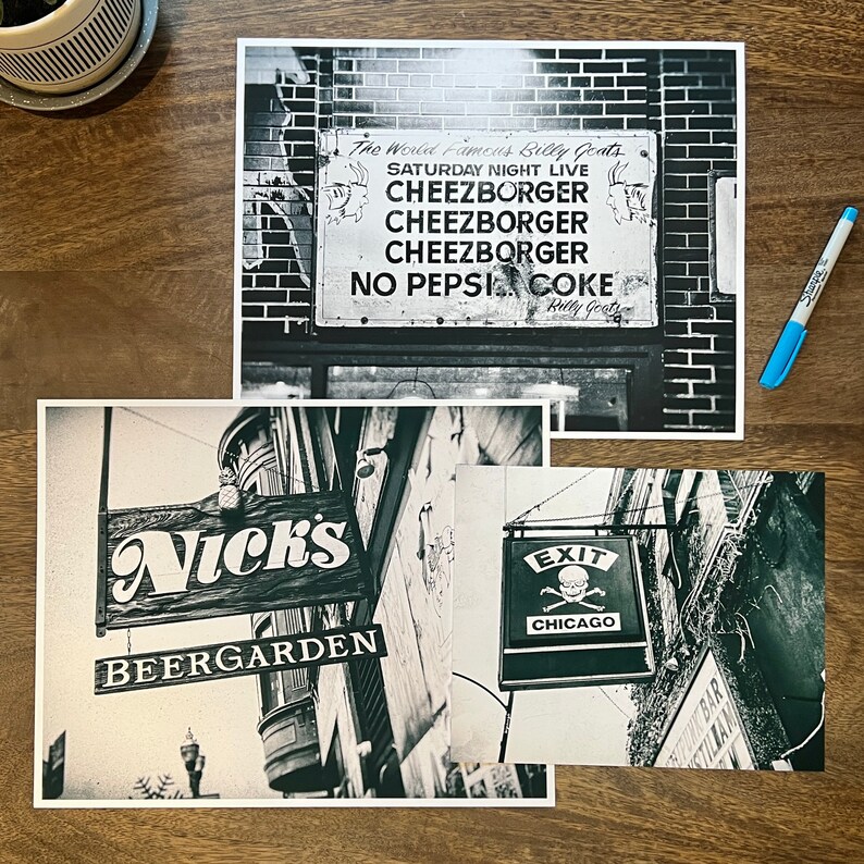 Op de afbeelding: Drie zwart-witfoto's van bezienswaardigheden in Chicago. De eerste foto toont een bord voor "The World Famous Billy Goats" met de tekst "Saturday Night Live, Cheezborger, Cheezborger, Cheezborger, No Pepsi Coke, Billy Goat". De tweede foto toont een bord voor "Nick's Beer Garden". De derde foto toont een bord met een schedel en gekruiste beenderen en de tekst "Exit Chicago".