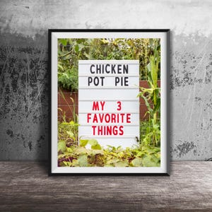May include: A framed sign with the words "CHICKEN POT PIE" and "MY 3 FAVORITE THINGS" in black and red lettering. The sign is set against a backdrop of greenery and a wooden surface.