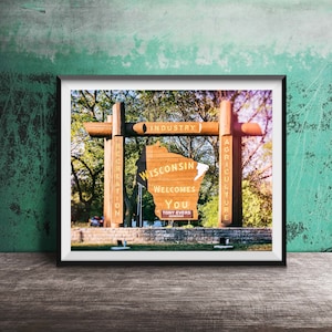 May include: A wooden sign shaped like the state of Wisconsin welcomes visitors with the text "Wisconsin Welcomes You" and the name of the governor, Tony Evers. The sign is surrounded by trees and a stone wall.
