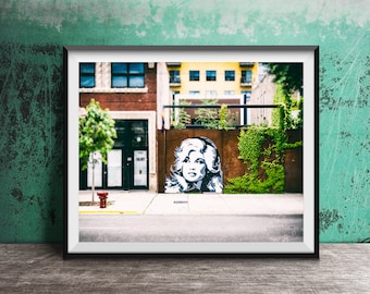May include: A framed wall art print featuring a street scene with a black and white mural of a woman with wavy hair painted on a rusted metal wall. The scene includes a brick building, a yellow building, a green vine, and a red fire hydrant.