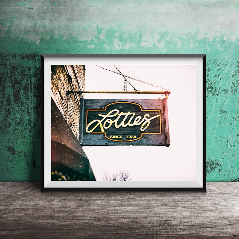 Pode incluir: Uma placa vintage com o texto "Lotties Since 1934" pendurada em um pr&eacute;dio. A placa &eacute; pintada de azul escuro com letras douradas. A placa est&aacute; pendurada em uma corrente e est&aacute; ligeiramente inclinada.
