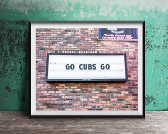 Może przedstawiać: Oprawiony wydruk przedstawiający ceglaną ścianę z napisem "GO CUBS GO" czarnymi literami. Znak jest podświetlony i umieszczony na tle czerwonych i brązowych cegieł. Wydruk jest oprawiony w czarną ramę i umieszczony na drewnianej powierzchni.