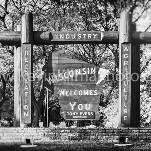 May include: Black and white photo of a wooden sign shaped like the state of Wisconsin. The sign reads "Wisconsin Welcomes You" and "Tony Evers Governor". The sign is surrounded by trees and a brick wall.