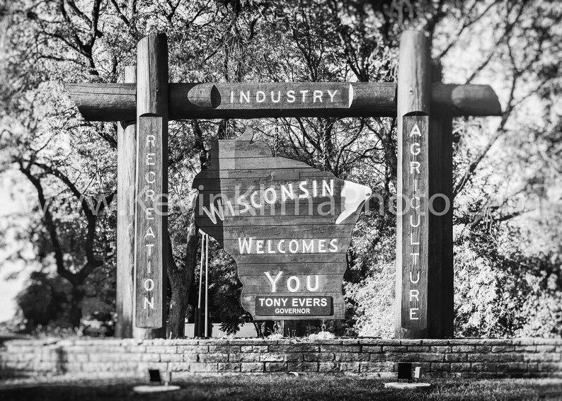 May include: Black and white photo of a wooden sign shaped like the state of Wisconsin. The sign reads "Wisconsin Welcomes You" and "Tony Evers Governor". The sign is surrounded by trees and a brick wall.