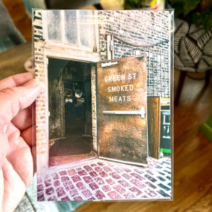 Może przedstawiać: Wizerunek sklepu w stylu vintage z drewnianymi drzwiami, na kt&oacute;rych widnieje napis "Green St Smoked Meats". Drzwi są otwarte, odsłaniając brukowaną ścieżkę prowadzącą do budynku.