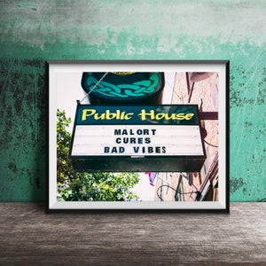 May include: A green and yellow sign with the words "Public House" and "Malort Cures Bad Vibes" hanging above a building.
