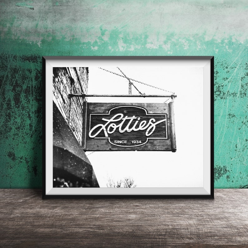 Pode incluir: Fotografia a preto e branco de uma placa pendurada num edif&iacute;cio que diz "Lotties Since 1934".