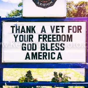May include: A sign in blue and white with the message "THANK A VET FOR YOUR FREEDOM GOD BLESS AMERICA" in black lettering. The sign is positioned outdoors, with a road and building visible.