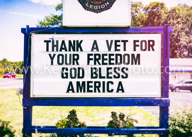 May include: A sign in blue and white with the message "THANK A VET FOR YOUR FREEDOM GOD BLESS AMERICA" in black lettering. The sign is positioned outdoors, with a road and building visible.